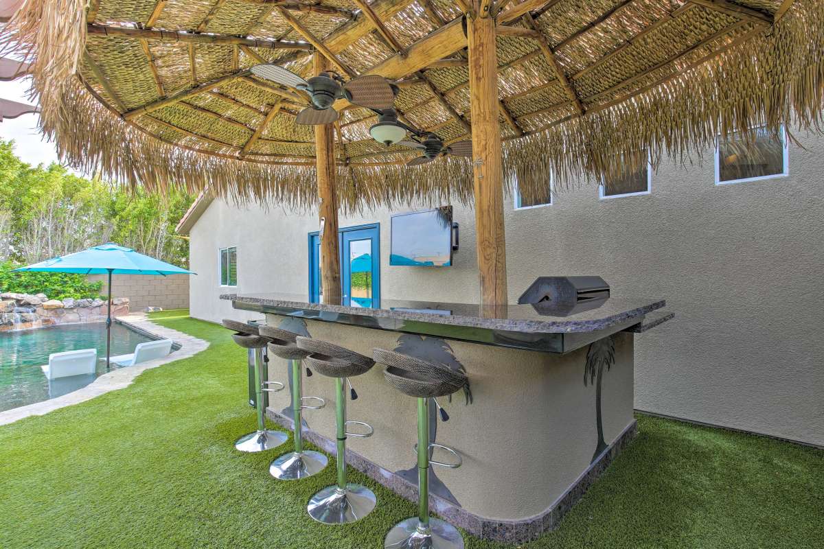An outdoor tiki bar features a thatched roof supported by wooden beams, equipped with a granite countertop and four bar stools. A 55" outdoor TV is mounted nearby, and a vibrant blue umbrella offers shade over the bar area, creating a relaxed setting by the pool.