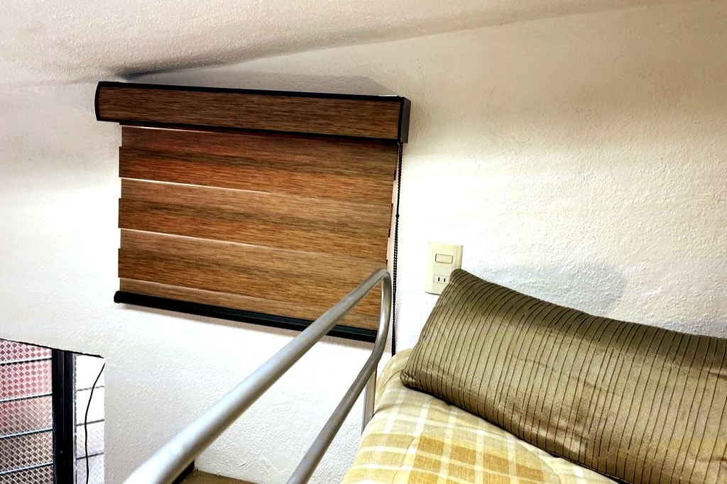 A corner of a cozy sleeping area features a bed with a striped pillow and a plaid blanket. A wooden blind is partially closed, allowing soft light to filter through, while a metal railing adds a touch of modern design.