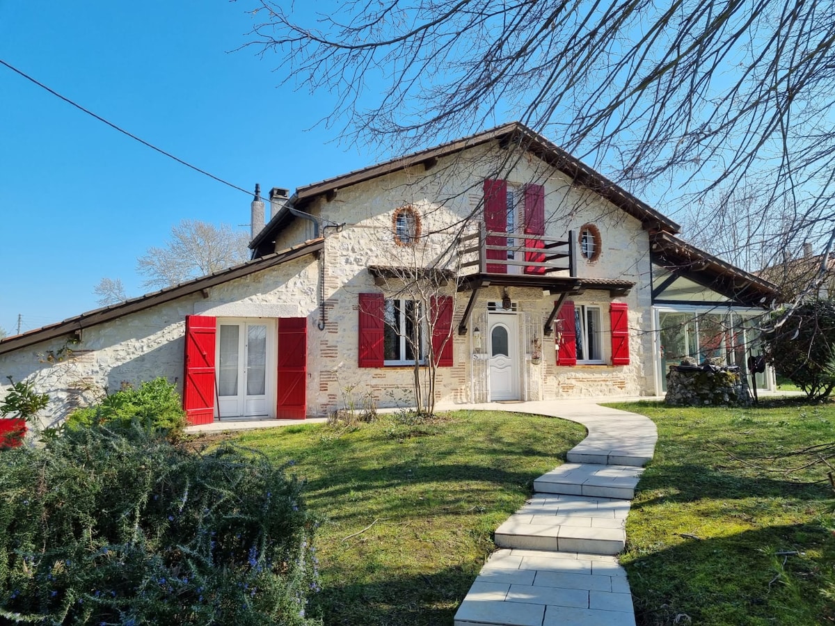 A renovated stone farmhouse stands amidst a spacious garden, featuring red shutters and a welcoming front pathway. Large windows invite natural light, while the scenic exterior is complemented by the surrounding greenery and a terrace balcony above the entrance.