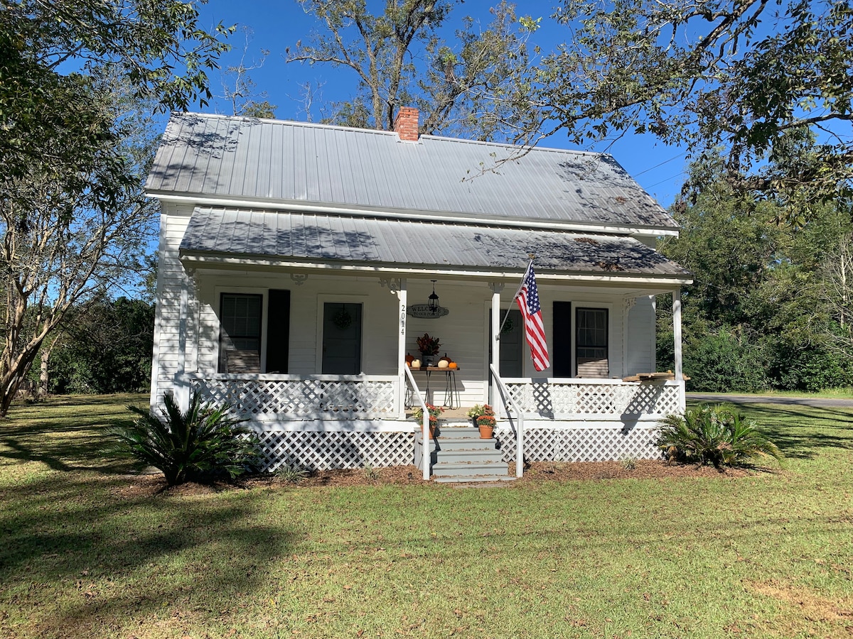 The charming cottage exterior features a metal roof and a welcoming porch adorned with potted plants. An American flag gently flutters in the breeze, while surrounding greenery and a spacious lawn add to the serene atmosphere.