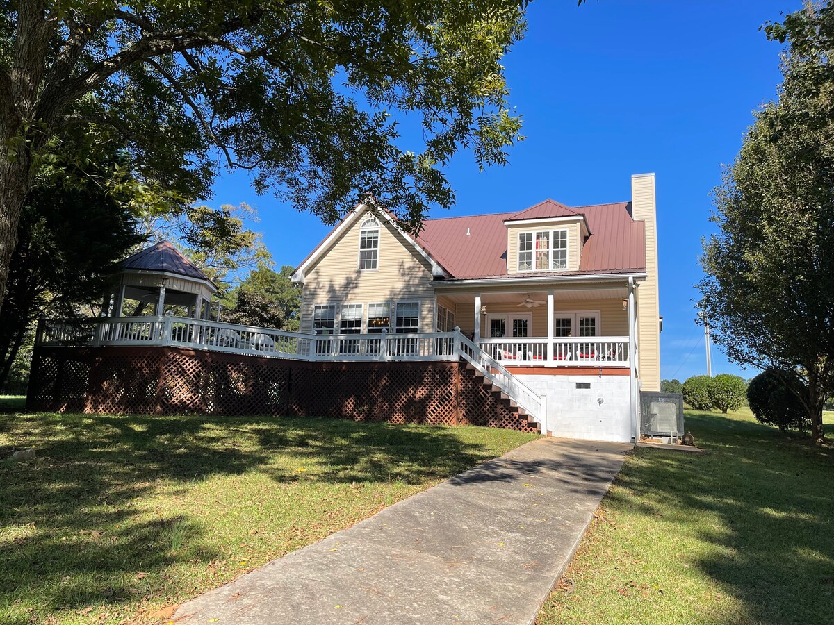 A spacious two-story home with a large front porch and a sloped driveway is showcased. Surrounding greenery and clear blue skies contribute to a welcoming outdoor environment. A gazebo is visible in the yard, complementing the tranquil setting.