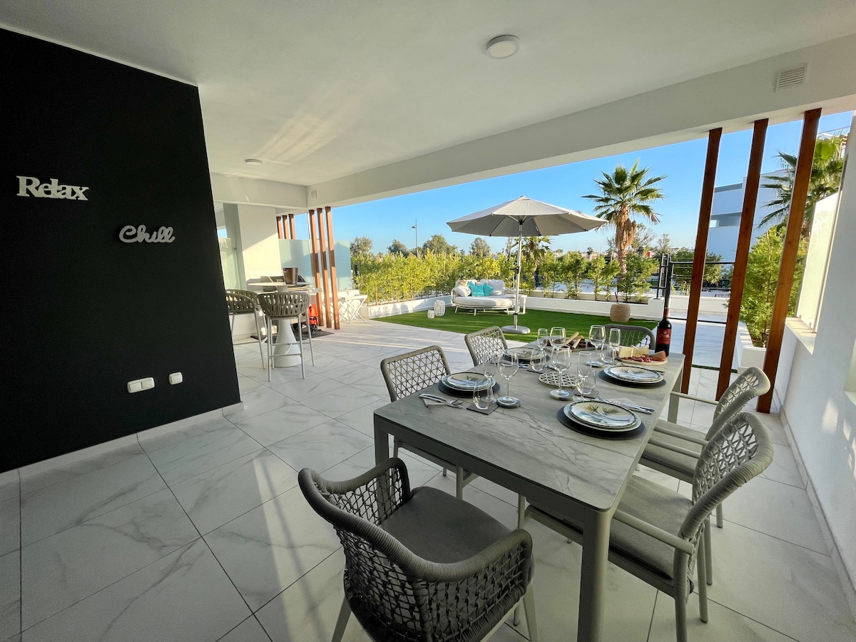 An outdoor dining area features a large table set for meals, complemented by stylish chairs. A patio umbrella provides shade, while lush greenery and palm trees create a serene atmosphere. Access to a pool is visible in the background, enhancing the space's tranquil feel.