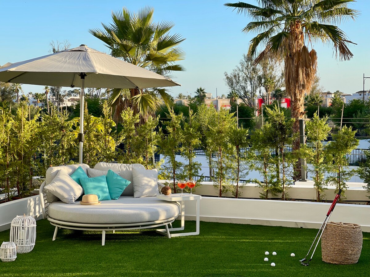 A cozy outdoor seating area features a circular lounge bed adorned with light blue cushions, positioned beneath a large umbrella. Lush greenery surrounds the space, providing a serene backdrop. A small putting green with golf balls is visible, enhancing the leisure atmosphere.