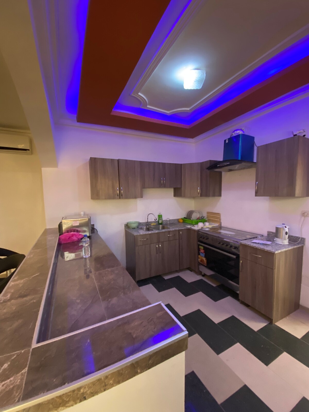 A contemporary kitchen is displayed, featuring dark wooden cabinets and a marble countertop. Modern appliances are present, including an oven and a refrigerator. Ambient lighting in blue casts a soft glow, complementing the contrasting black and white tiled flooring.