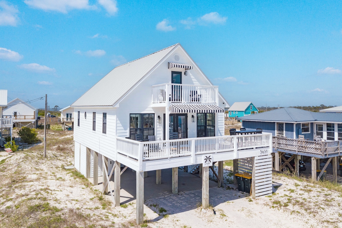 The exterior of a two-story beach house features a light-colored siding and a metal roof. A spacious deck with chairs overlooks the surrounding landscape. The structure is elevated on stilts, providing an unobstructed view of the nearby sandy area and homes.