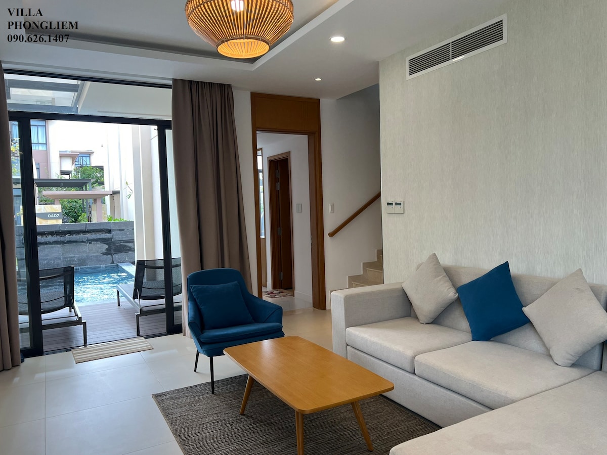 A spacious living area features a light-colored sofa with blue accent pillows, complemented by a wooden coffee table. Large glass doors open to a private pool area, while natural light brightens the room. Neutral walls and flooring enhance the modern design.