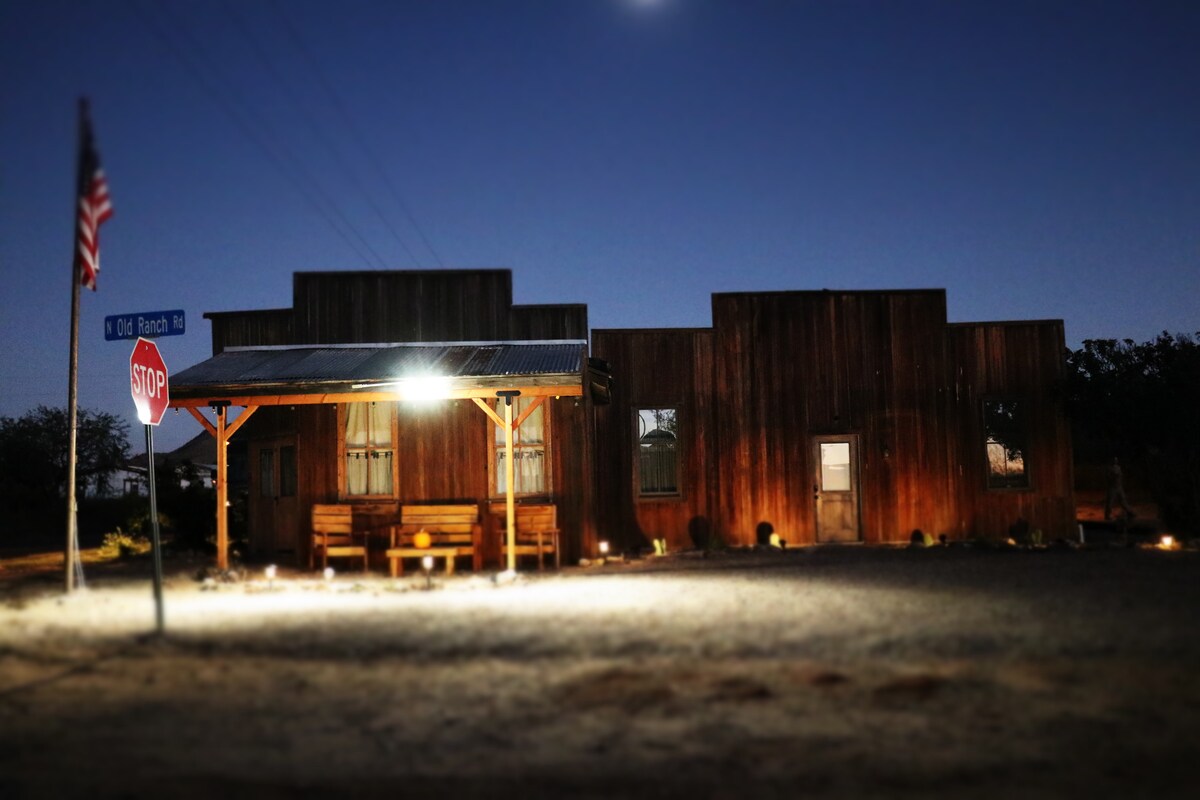 The exterior of the historic JH Smith building is illuminated at night. A wooden facade features distinct architectural lines, complemented by a covered porch and two benches. A stop sign stands nearby, with subtle lighting enhancing the rustic charm of the structure.