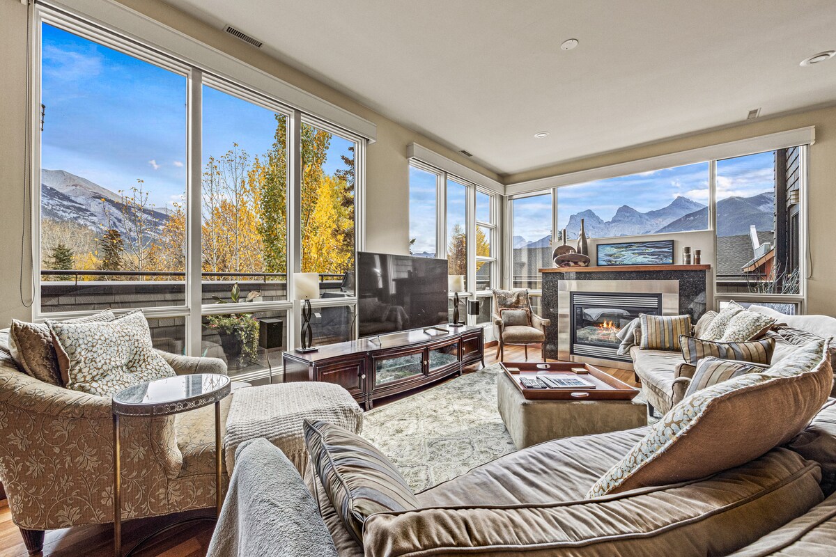 A spacious living area features large windows that offer views of the surrounding mountains and autumn foliage. Comfortable seating arrangements include plush armchairs and a sofa, while a modern fireplace adds a cozy focal point. A television is positioned on an elegant wood console.