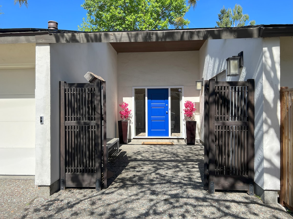 The entrance is framed by two dark wooden gates, leading to a vibrant blue front door. Two large planters filled with pink flowers are positioned on either side, enhancing the inviting feel of the pathway that is surrounded by greenery.