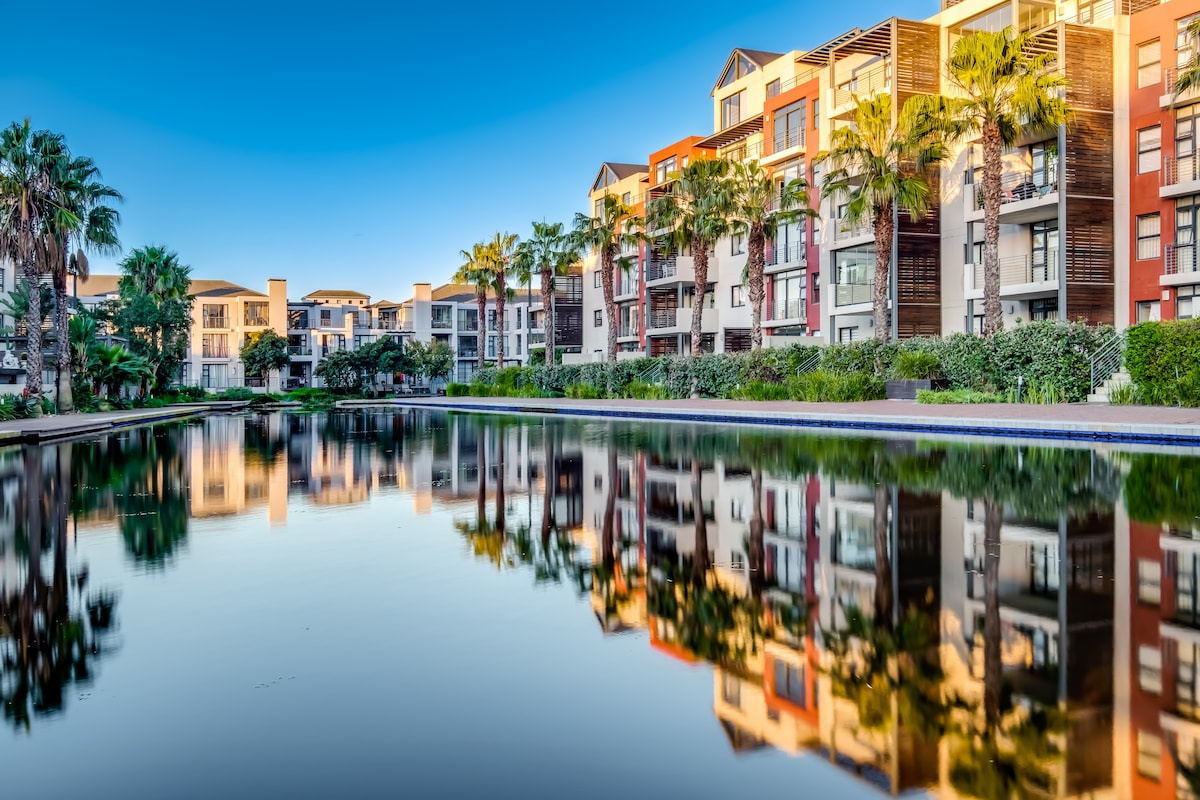 A tranquil canal reflects the surrounding apartment buildings, which feature a mix of colorful facades and greenery. Palm trees line the water's edge, providing a sense of serenity and enhancing the natural beauty of the environment.