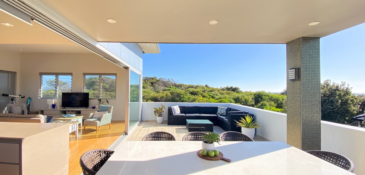 An open-air balcony presents a spacious seating area with comfortable outdoor furniture, surrounded by lush greenery and expansive views of the landscape. Large glass doors connect the balcony to an interior living space, allowing natural light to illuminate the area.