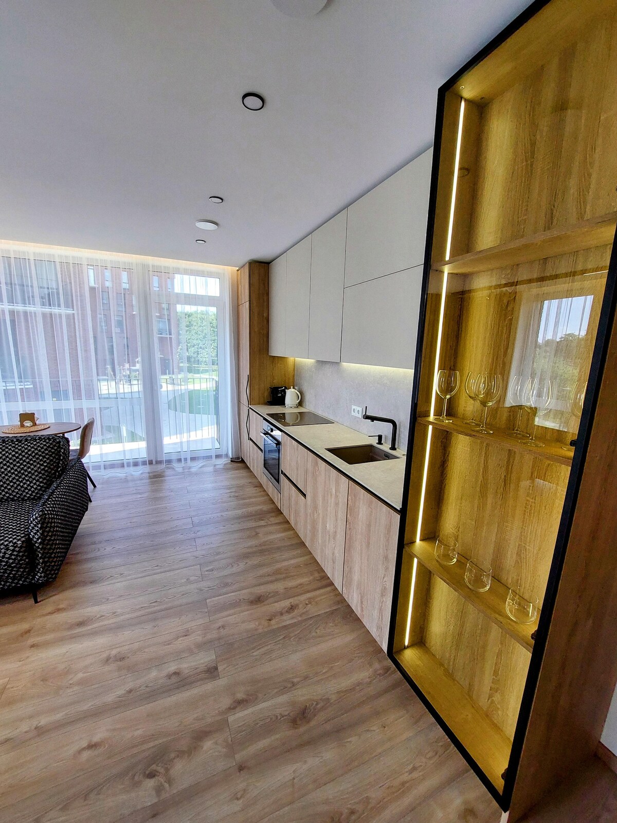 A modern kitchen area features sleek cabinetry with wood accents and a marble countertop. Glassware is displayed on illuminated shelves. Large windows allow natural light to fill the space, enhancing the airy feel of the room.