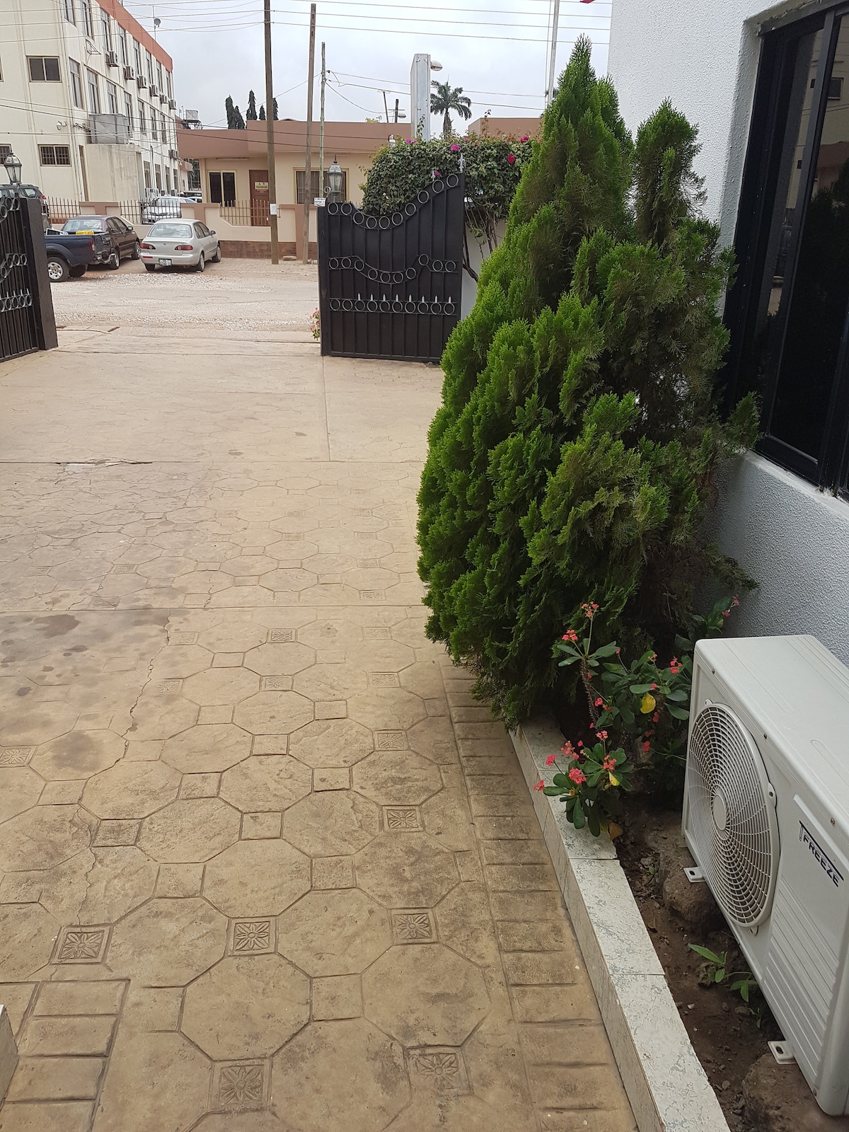 A well-maintained entrance area is depicted, featuring a patterned concrete pathway leading towards a gate. Lush greenery, including a bush and flowering plants, is visible beside an air conditioning unit, adding a touch of nature to the surroundings.