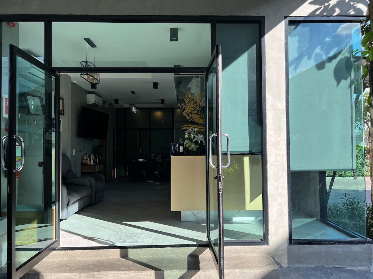 The entrance features large glass doors that provide a clear view of the reception area. Natural light floods the space, reflecting off the modern furnishings and decor. Potted plants are positioned around the entrance, adding a touch of greenery to the contemporary design.