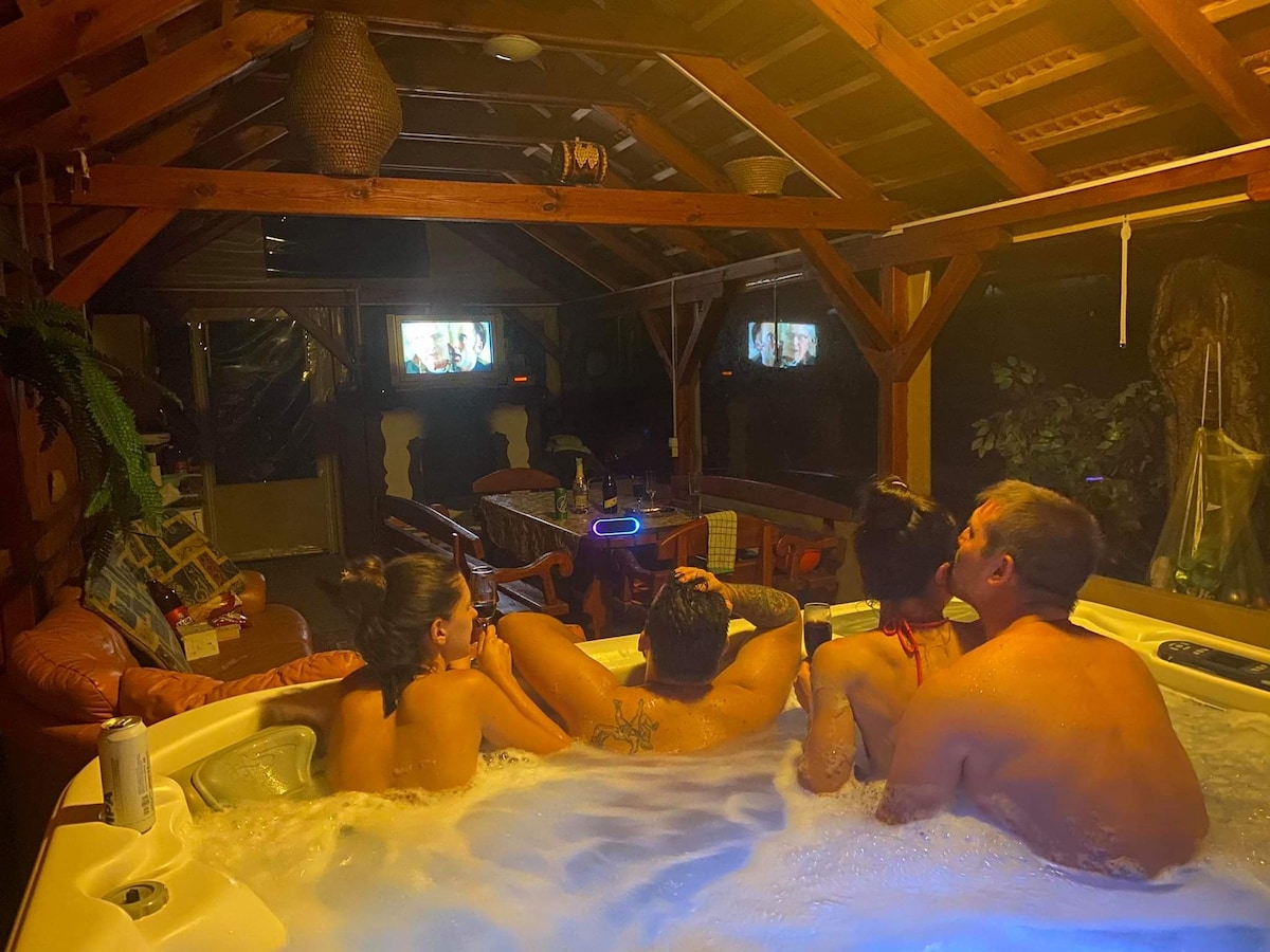 An interior space features a spacious hot tub filled with guests enjoying a relaxed atmosphere. Surrounding the tub, comfortable seating and cozy decor are visible. A TV is mounted on the wall, providing entertainment in the background, while soft lighting enhances the warm ambiance.