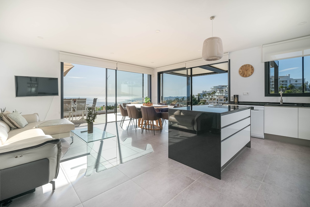 A modern open-plan living area is showcased, featuring a kitchen island with a sleek black finish. Large glass doors provide panoramic views of the Mediterranean, while a spacious dining table and comfortable seating area create an inviting space filled with natural light.