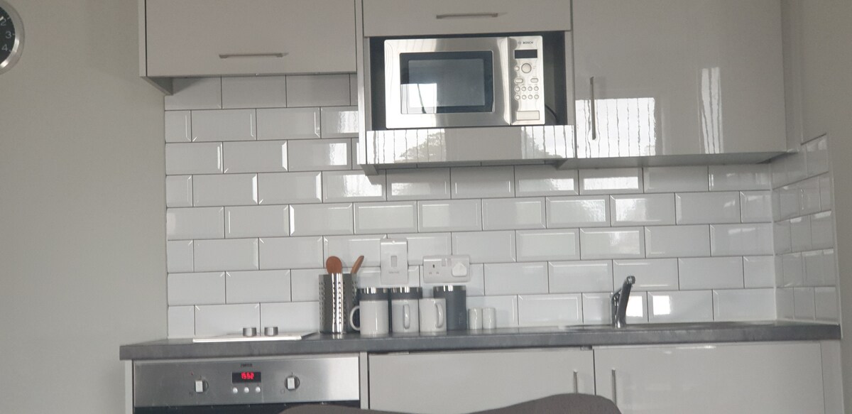 A modern kitchen area features sleek grey cabinets and a light-colored backsplash of glossy tiles. A microwave is mounted above a countertop equipped with cooking appliances and utensils. Natural light reflects off the surfaces, enhancing the clean design of the space.