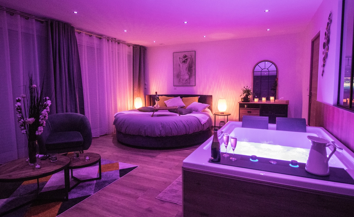 A spacious room features a round bed with soft bedding, accented by mood lighting in purple tones. A large jetted tub is placed nearby, accompanied by elegant decor. Candlelight creates a warm atmosphere, complemented by a small seating area with a modern chair and table.