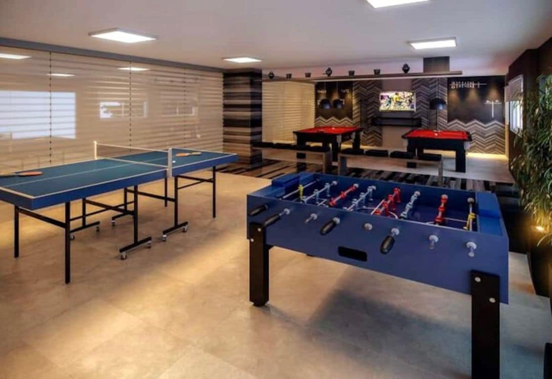 A recreational area is equipped with several game tables, including ping pong and foosball. A large television is mounted on the wall, and two billiard tables are positioned nearby. Natural light filters in through the window blinds, creating a bright and open atmosphere.