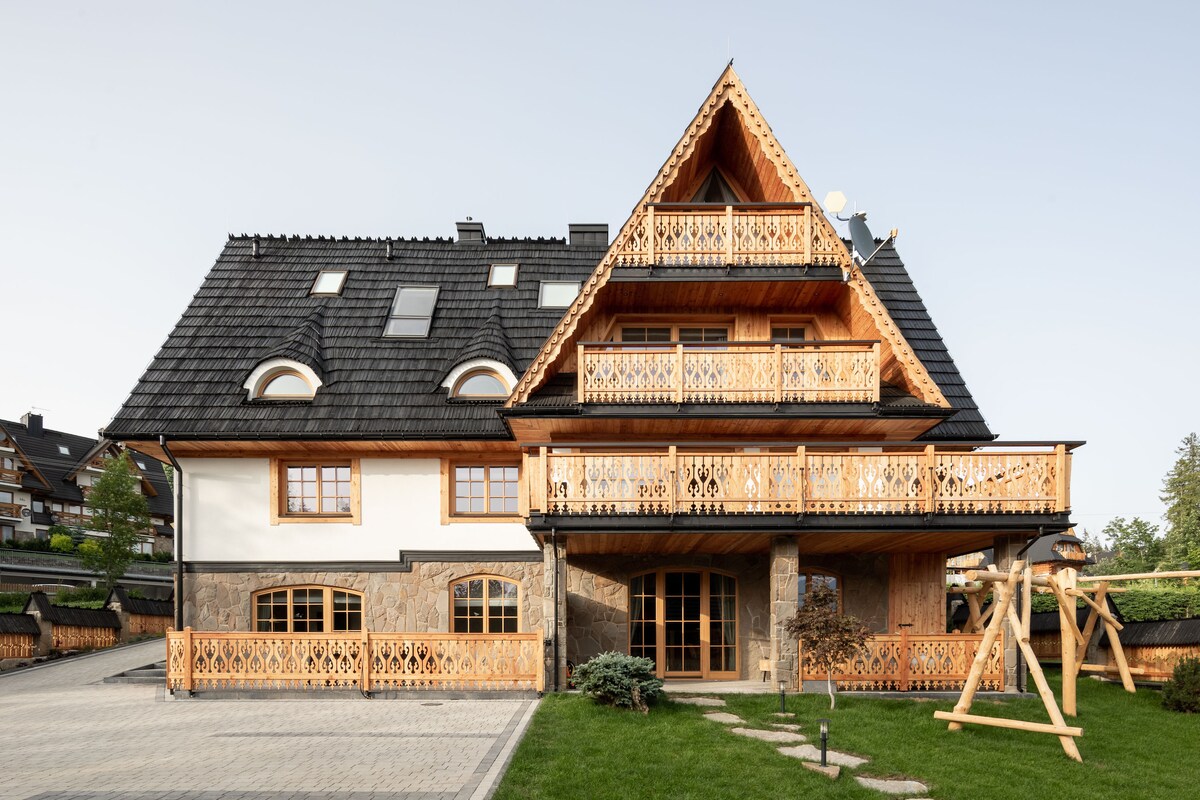 The exterior of a stylish apartment complex is displayed, featuring a blend of wooden and stone elements. Balconies with intricate wooden railings overlook the landscaped yard. A swing is positioned on the grass, and a paved area provides access to the entrance.