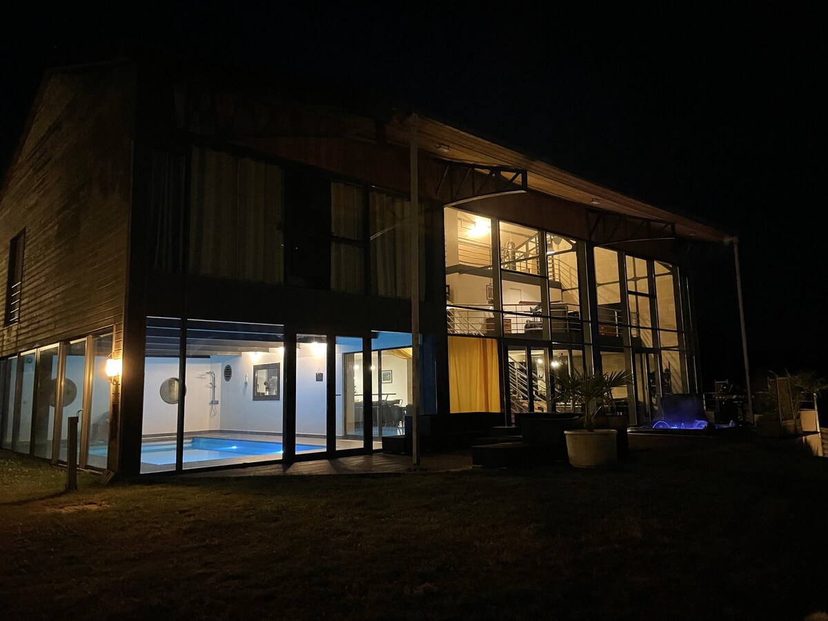 The villa's exterior is illuminated at night, showcasing large glass panels that reflect the surroundings. A bright blue indoor pool is visible through the windows. The structure combines industrial and marine designs, surrounded by green grass and a minimalist outdoor area.