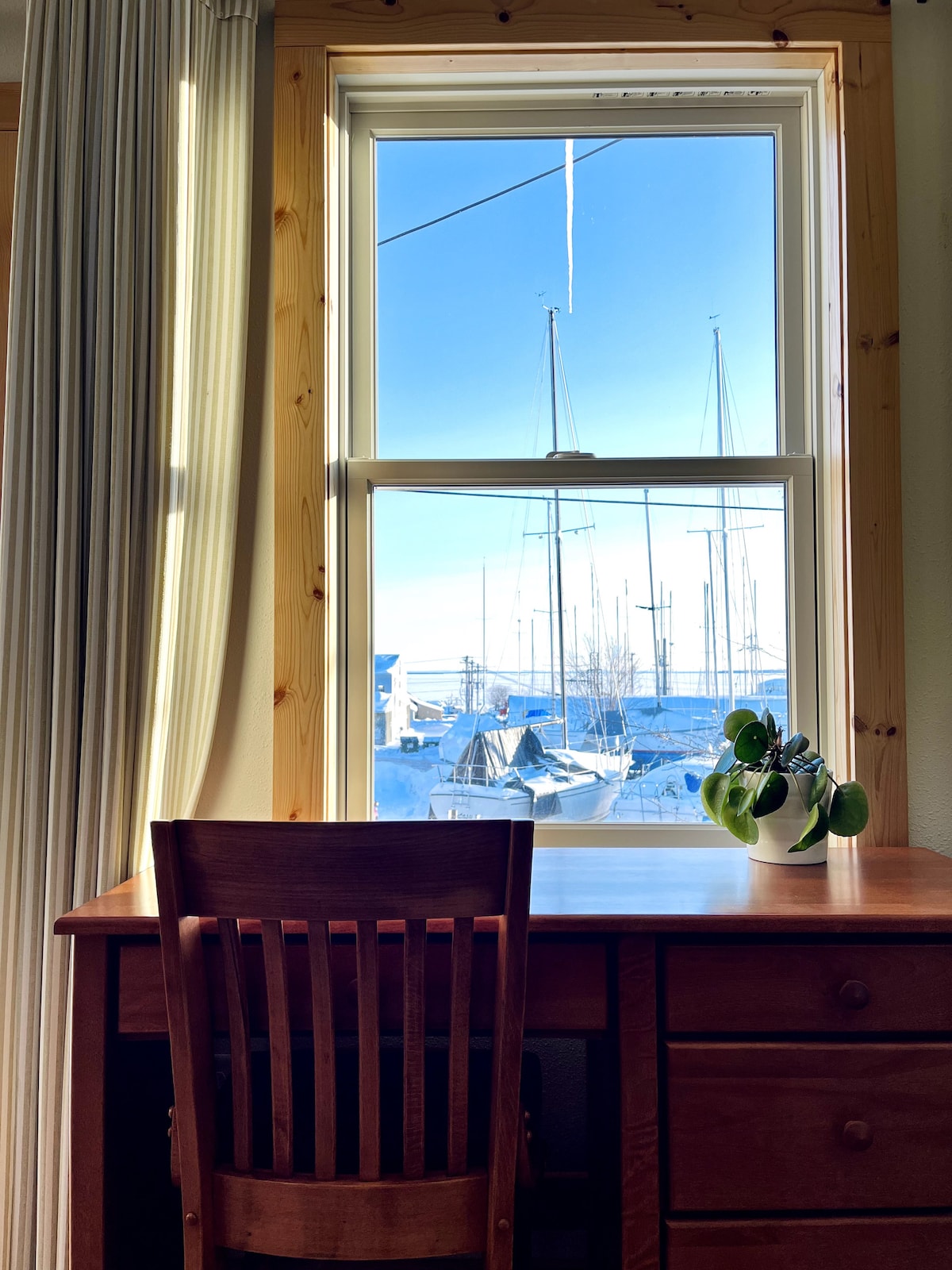 A wooden desk with a chair is positioned near a window that offers a view of boats docked in the marina. The south-facing window allows natural light to illuminate the space, with light curtains framing the view.
