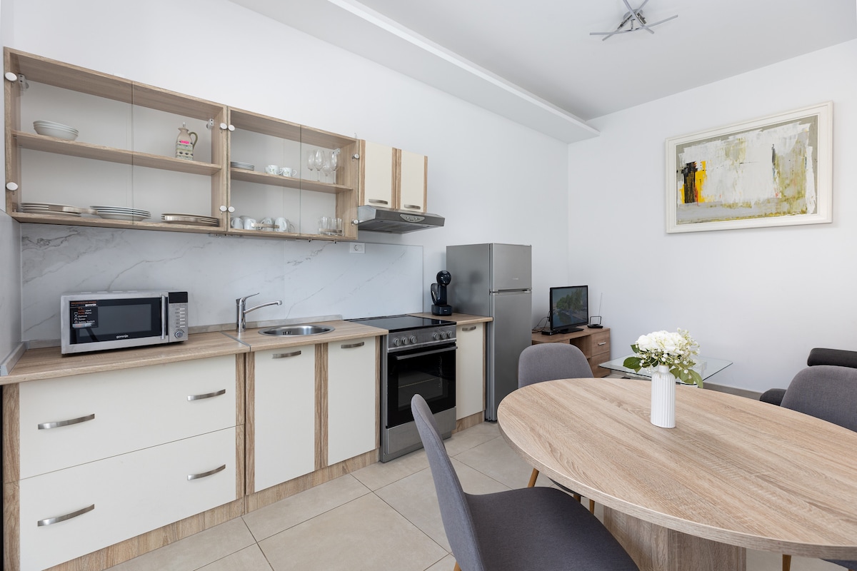 The modern kitchen showcases sleek cabinetry paired with a smooth countertop. Essential appliances, including a microwave, stove, and refrigerator, are neatly arranged. A round dining table is positioned nearby, surrounded by gray chairs, creating a functional dining space.