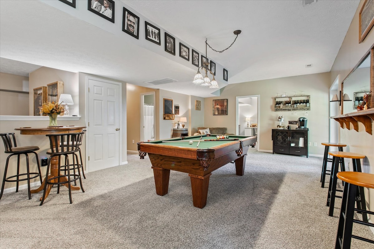 A spacious recreational area is highlighted by a large pool table at the center. A bar area with high stools complements the setup. The walls are decorated with framed photographs, and soft lighting enhances the inviting ambiance. A coffee station is visible in the background.