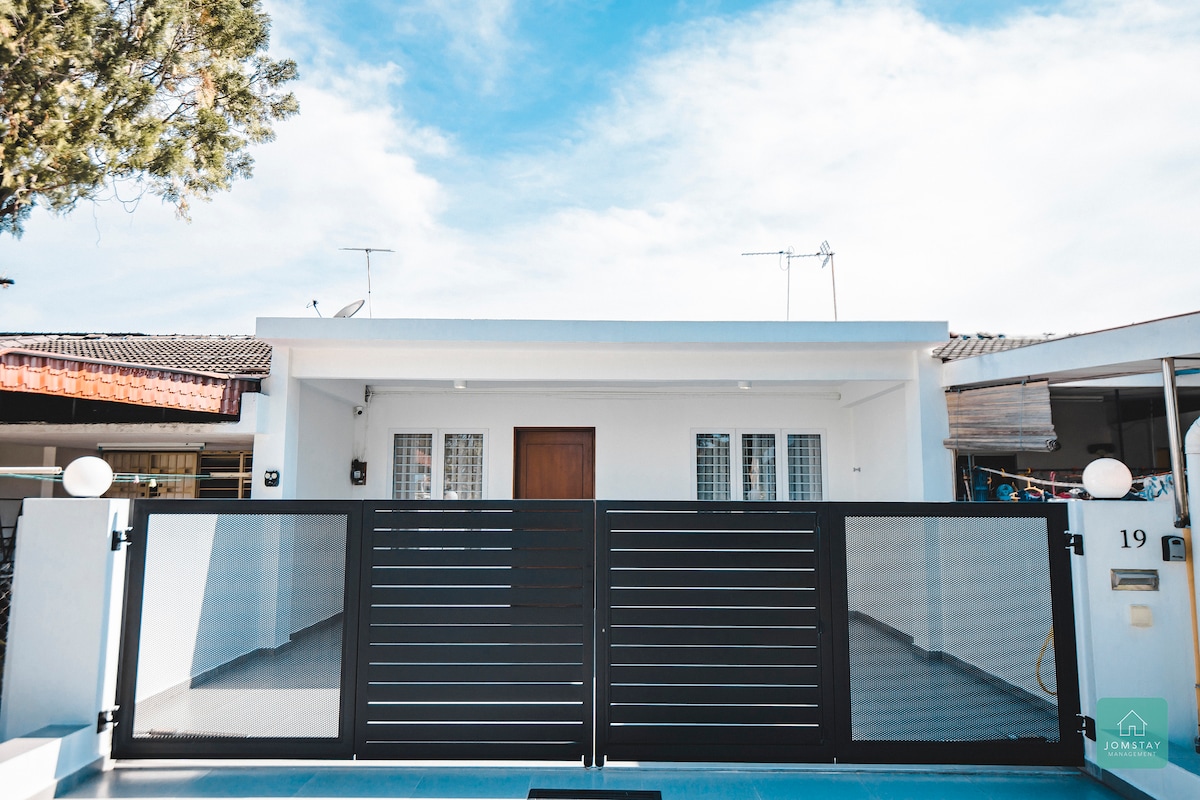 The front view of the single-storey house displays a modern facade with a simple, clean design. A black metal gate leads to a spacious entryway, with large windows allowing natural light to fill the interior. The exterior is painted in bright neutral tones.