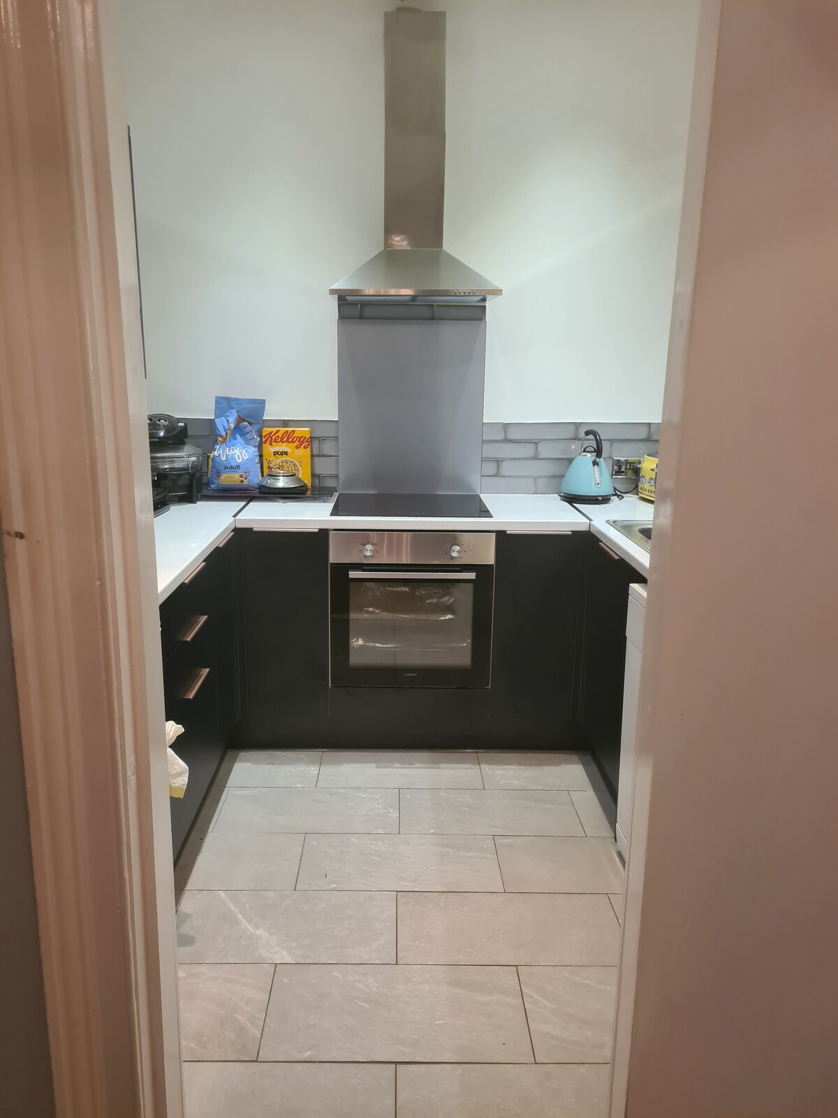 The kitchen features modern cabinetry in dark tones, with a sleek silver oven and a range hood above. Light grey tiled flooring complements the space, while smaller kitchen appliances, including a kettle, are visible on the countertop.