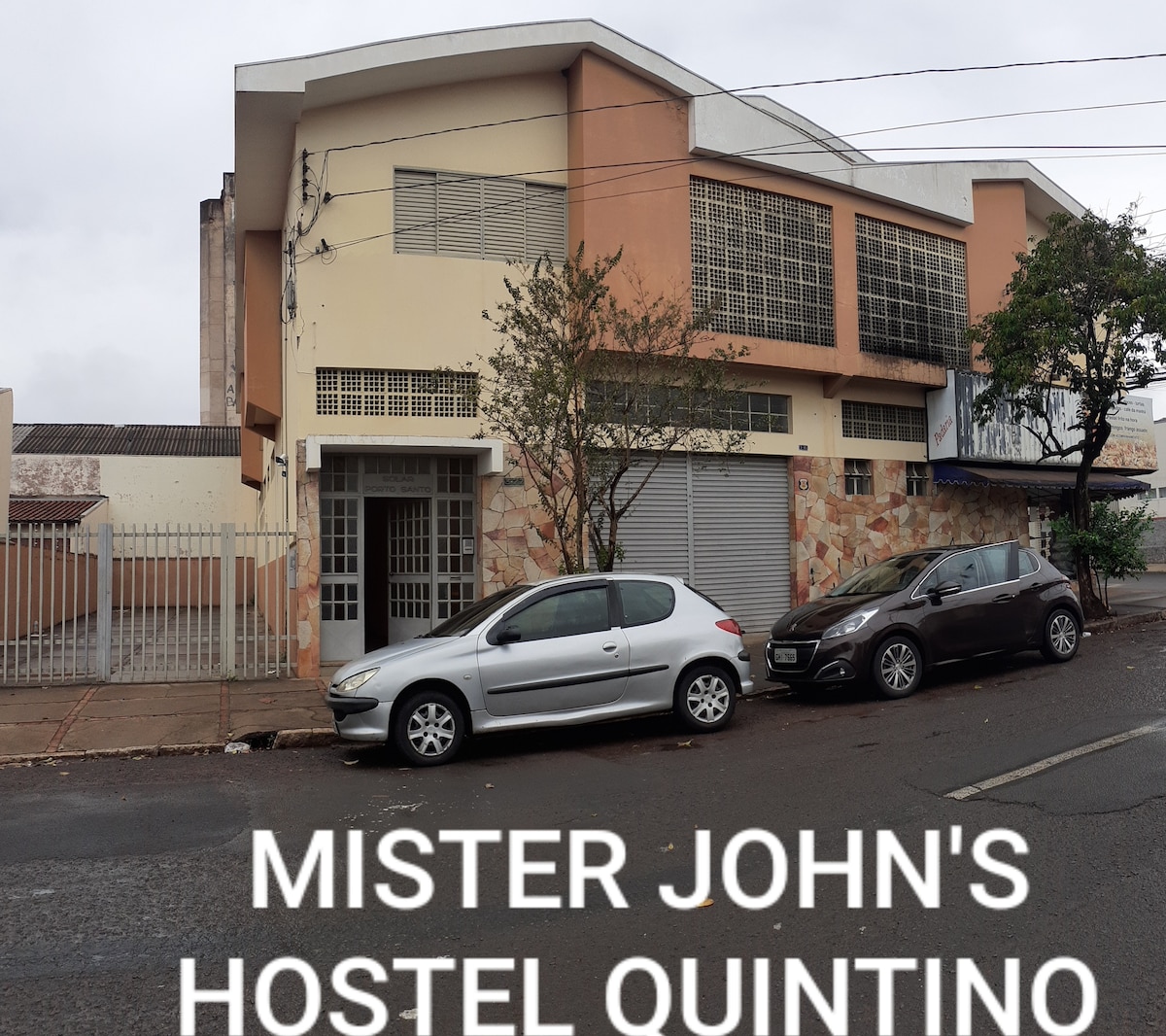 A large hostel building features a modern facade with a blend of stone and concrete materials. The entrance is framed by glass windows, and trees are positioned along the sidewalk. Two parked cars are visible in front, creating a welcoming environment.