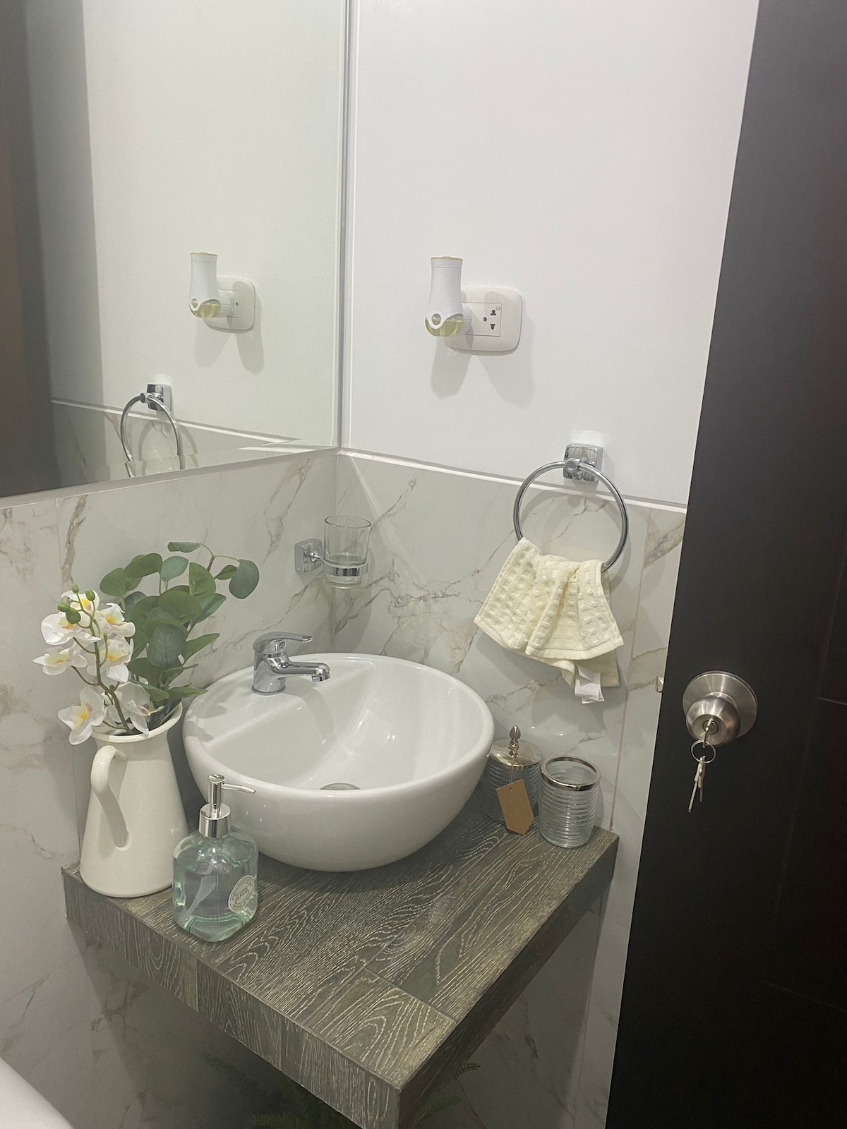 A modern bathroom corner is featured, displaying a vessel sink atop a wooden shelf. An orchid plant and decorative items adorn the area. A towel and soap dispenser are conveniently placed nearby, while a mirror reflects the space, enhancing the sense of openness.