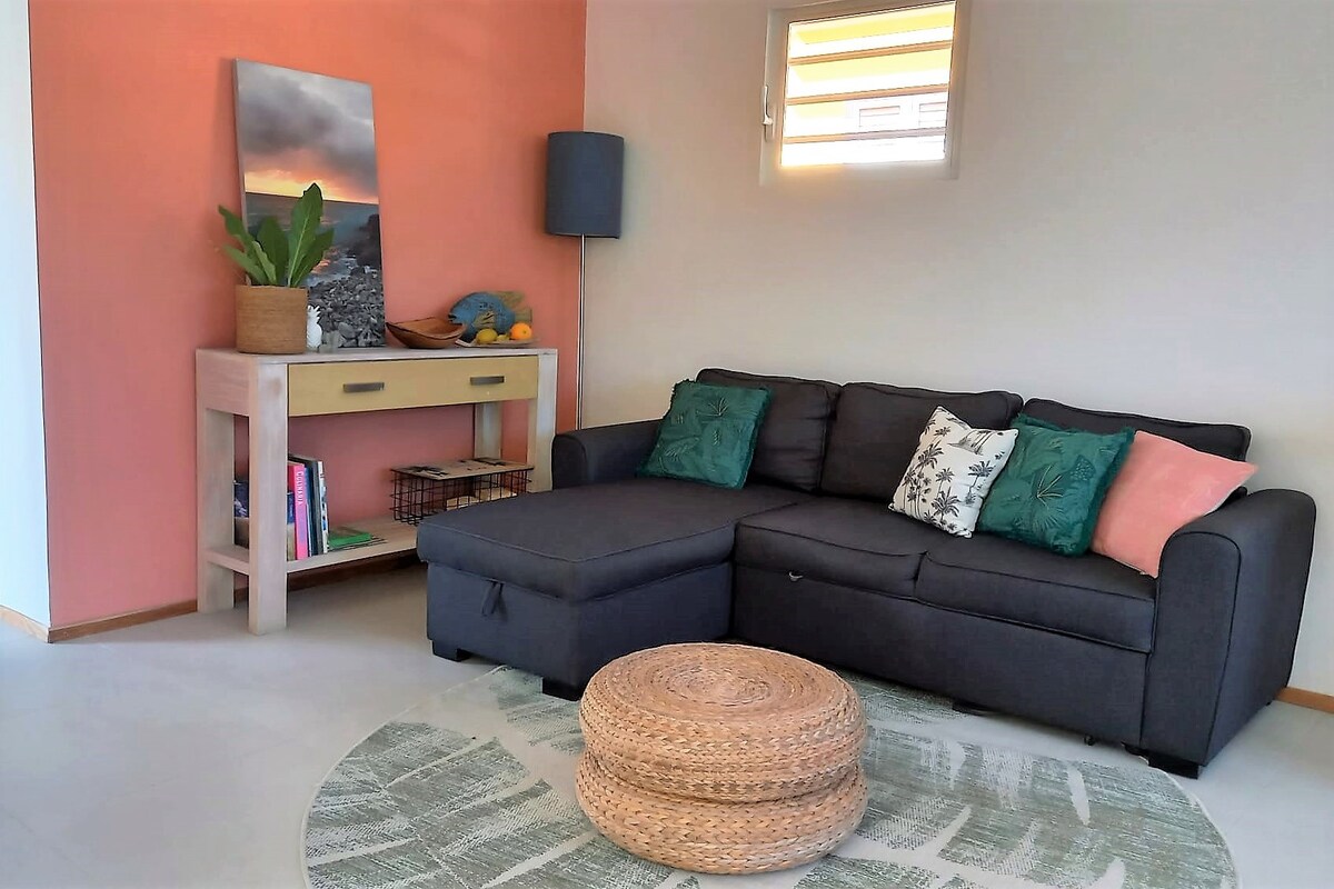 A cozy living area is presented, featuring a dark sectional sofa adorned with cushions in shades of green and pink. A circular woven pouf sits on a patterned rug. A side table holds decorative items, while a wall-mounted lamp and a small potted plant add touches of warmth.