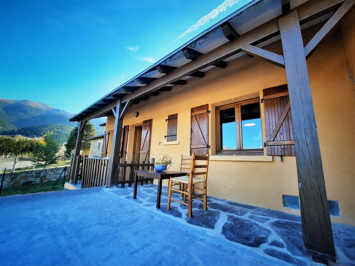 An inviting front terrace is shown, featuring a small table and a wooden chair. The stone flooring complements the warm exterior of the house, while wooden shutters adorn the windows. Scenic mountain views are visible in the background under a clear blue sky.