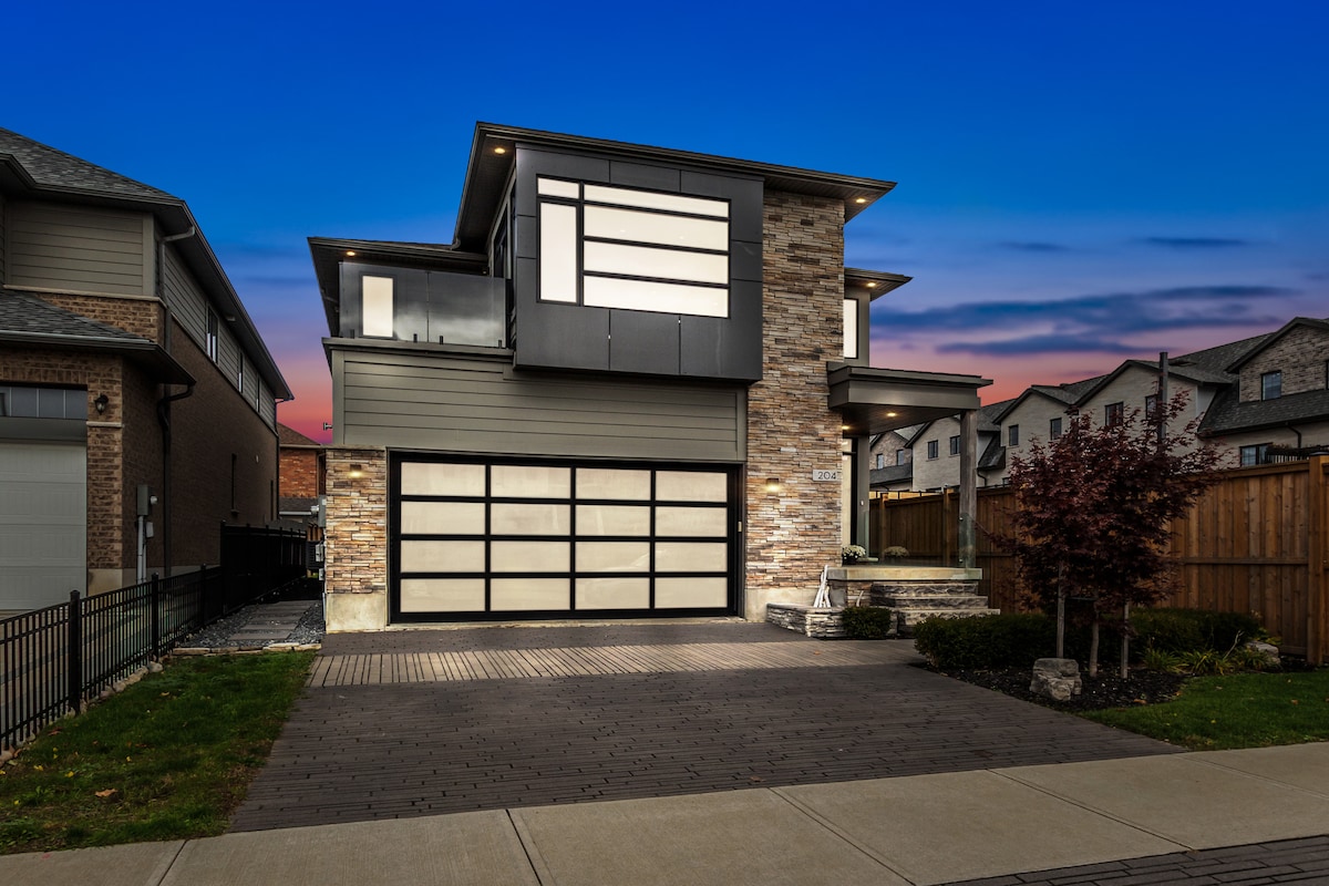 The modern two-story home features a striking façade of stone and wood, illuminated against a twilight sky. A large glass garage door complements the design, while neatly arranged landscaping and an inviting entryway enhance the overall aesthetic.