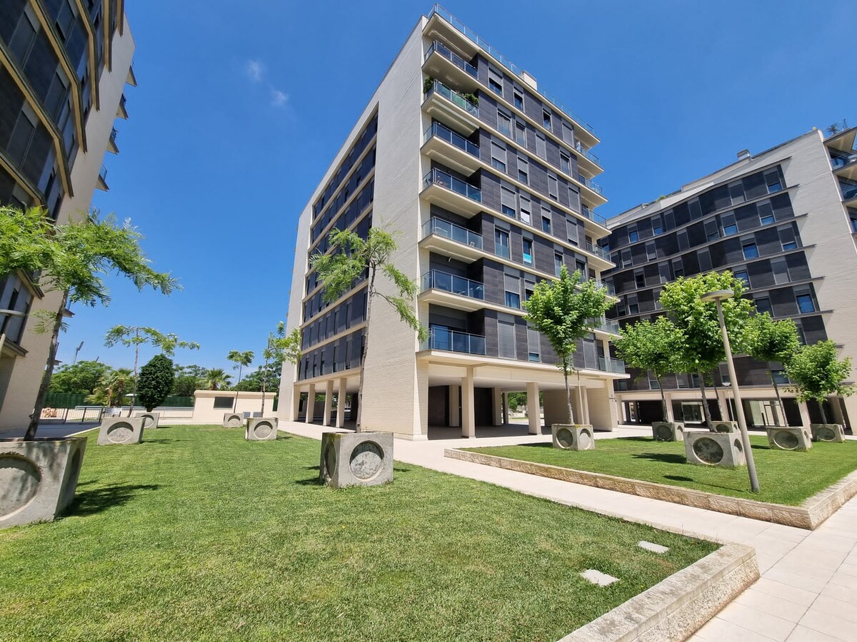 A modern apartment building is surrounded by well-maintained green lawns and strategically placed trees. The structure features multiple floors with large windows and balconies, contributing to an open atmosphere. Paved pathways lead through the landscaped area, enhancing accessibility.