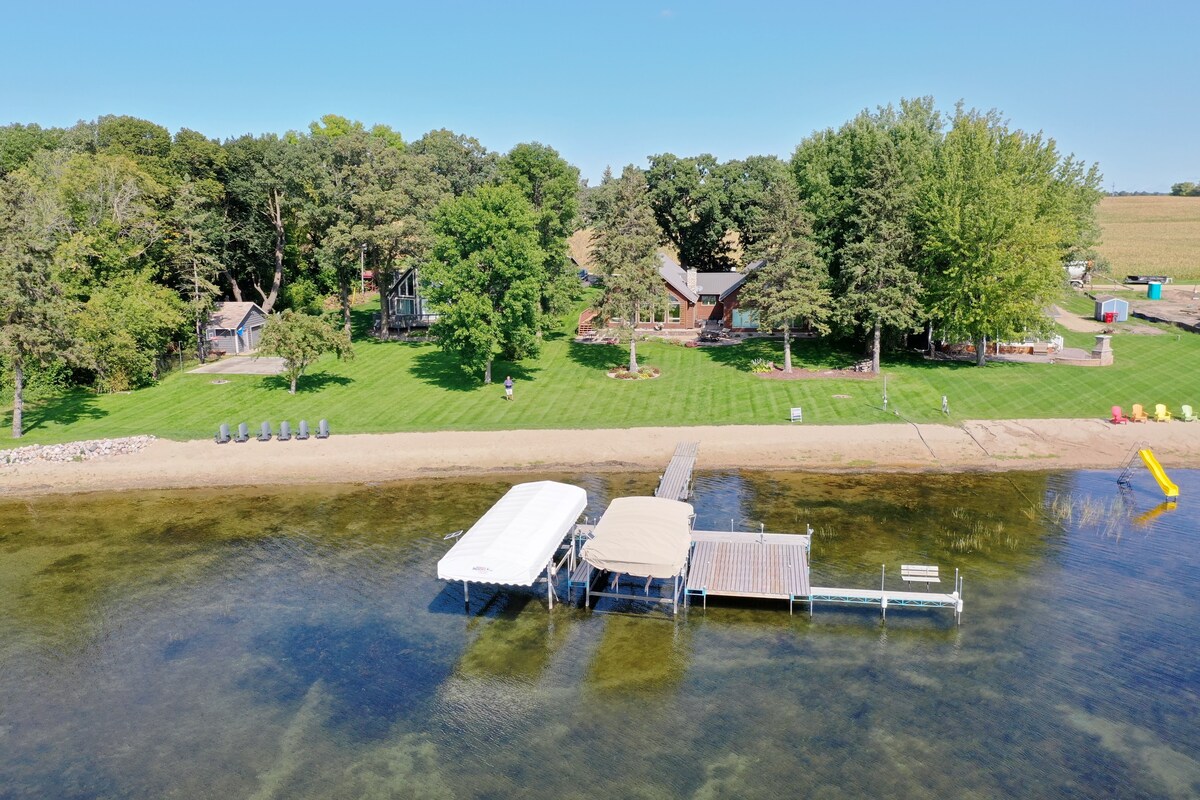 Aerial view of a spacious lakefront property featuring a sandy beach and dock. The large yard is visible with green trees lining the space. Two boat lifts are positioned on the dock, and vibrant outdoor equipment is arranged along the shore, enhancing the inviting lakeside setting.