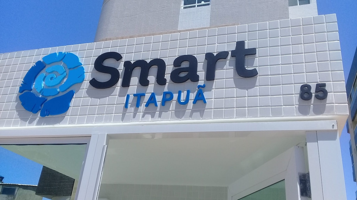A sign with the name 'Smart Itapuã' is prominently displayed on a light-colored tiled wall. The sign features a decorative blue swirl logo beside the text, and the number '85' is visible below it. Bright sunlight highlights the clean and modern appearance of the entrance.