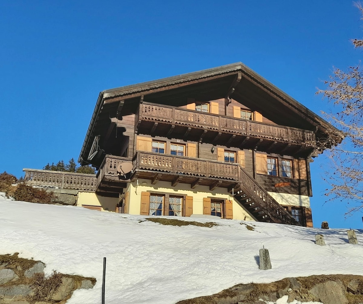 A traditional chalet is situated on a snowy slope under a clear blue sky. The two-story structure features wooden balconies with ornate railings and a combination of light and dark wooden siding. Steps lead up to the entrance, surrounded by a winter landscape.