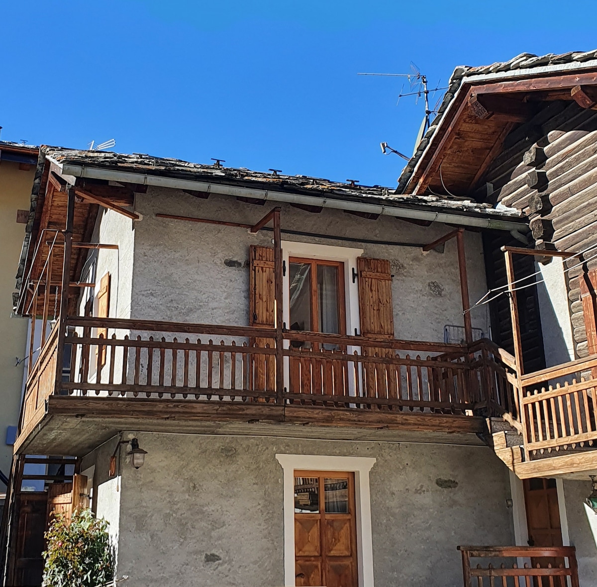 A charming mountain house exterior is presented, showcasing a wooden balcony with a railing. The facade features light grey walls and wooden shutters. The roof is tiled, and the entrance is marked by a wooden door at the base, surrounded by a small garden.