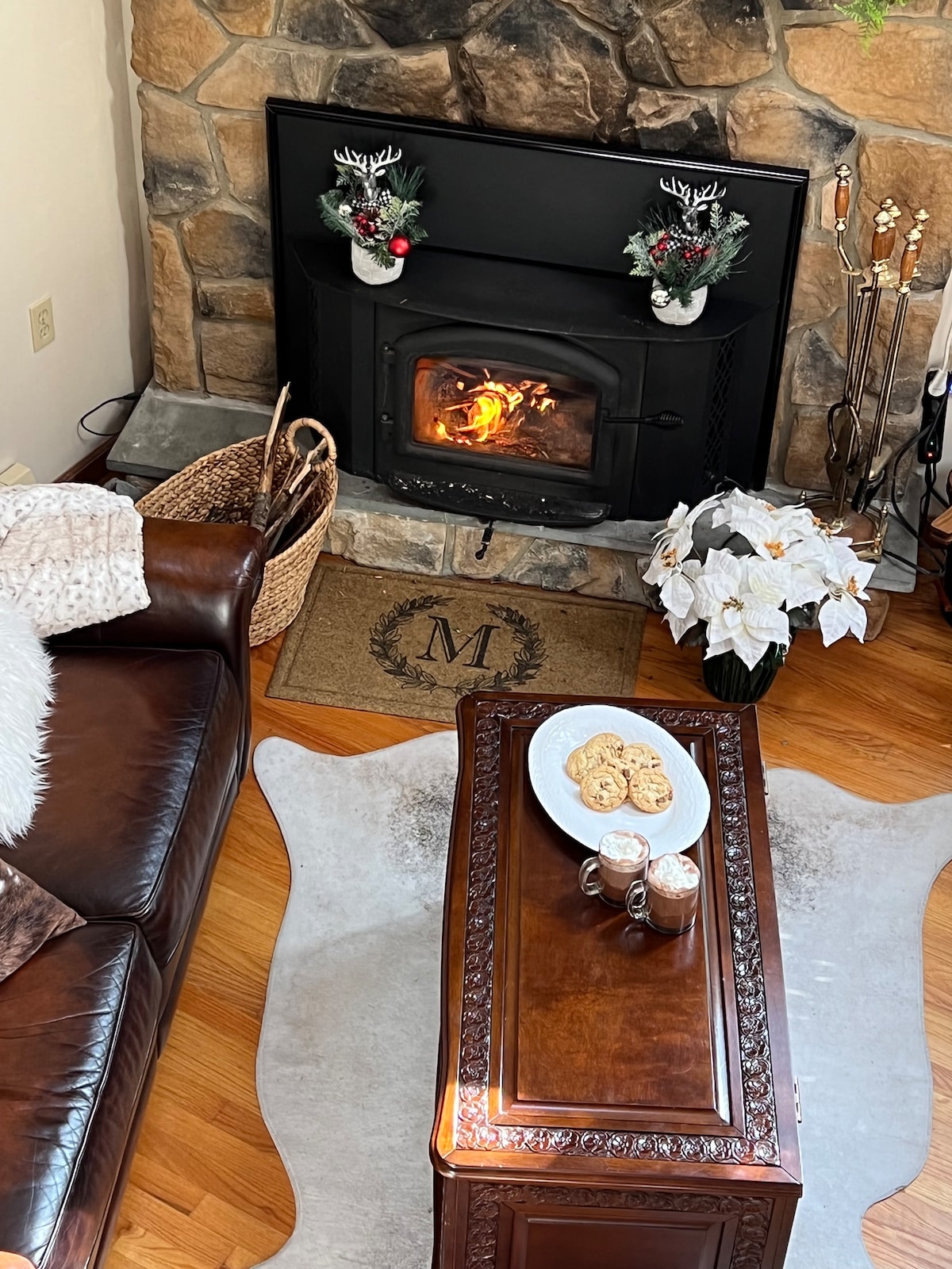A warm living area is highlighted by a stone fireplace, which showcases a gentle flame. A rustic coffee table sits in front, displaying a plate of cookies and two mugs. A plush brown sofa and a decorative basket add to the cozy ambiance.