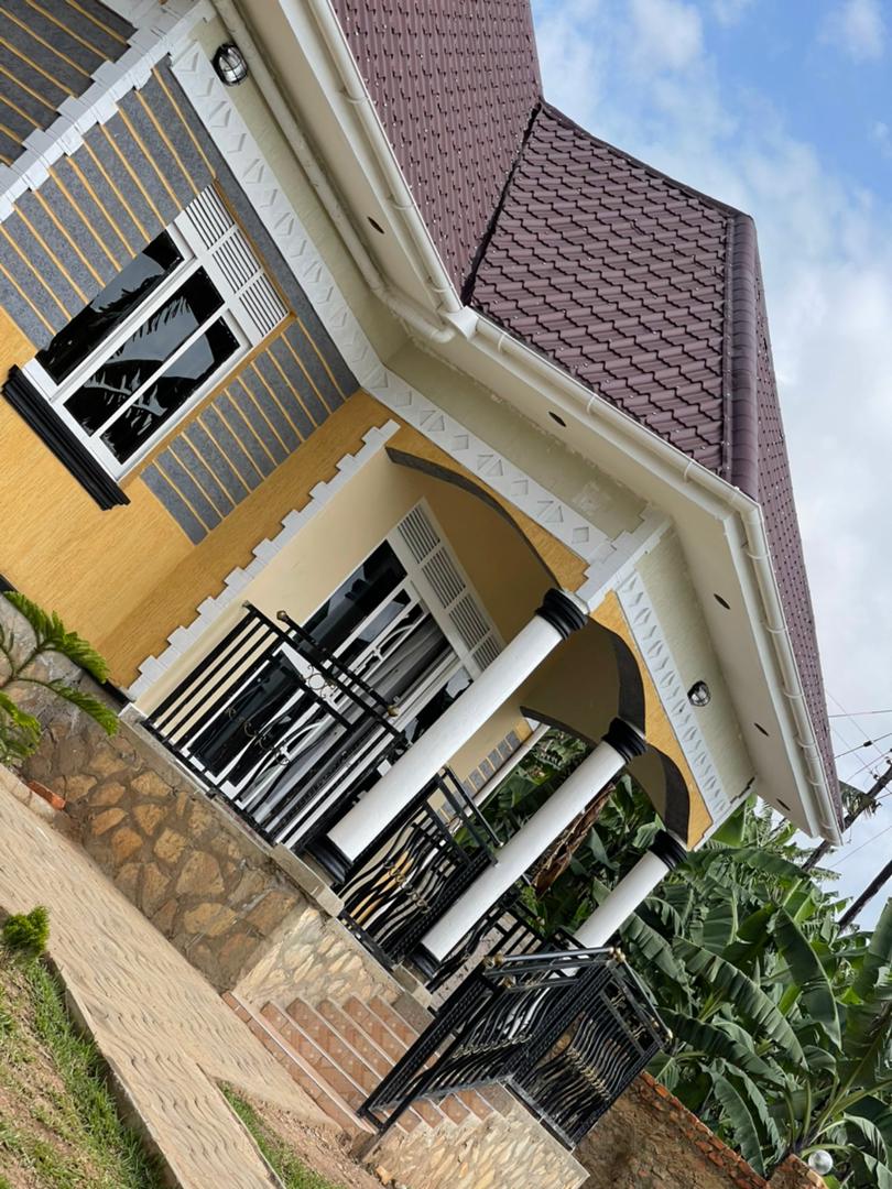 A unique single-story home is showcased with a sloped, dark roof and a mix of yellow and grey exterior walls. The porch is supported by elegant white columns, and large windows allow natural light to fill the interior. Lush greenery surrounds the property.