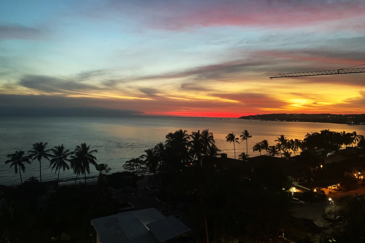 An expansive view of the ocean at sunset is captured, with vibrant colors blending in the sky. Silhouetted palm trees line the foreground, while distant lights from the shoreline reflect on the gentle water surface, creating a serene atmosphere.