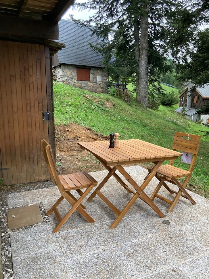 An outdoor patio area features a wooden table and two folding chairs, set against a backdrop of grass and trees. A small wooden storage unit is visible, and the rugged terrain leads toward additional chalet structures in the distance.