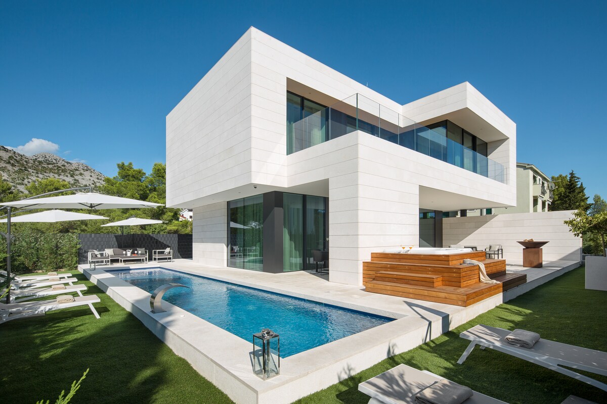 The modern villa showcases a clean architectural design with large glass windows. A private pool is featured in front, surrounded by a lush green lawn and sun loungers. An outdoor seating area is positioned next to the pool, providing a space for relaxation.