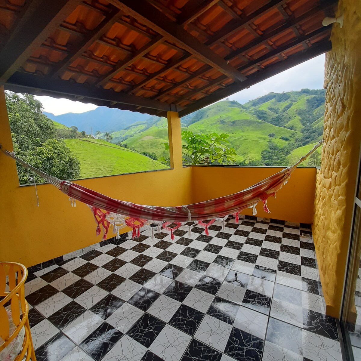 A spacious covered balcony features a vibrant hammock suspended between two supports. Black and white checkered tiles cover the floor, and large windows frame views of the surrounding green hills under a cloudy sky.