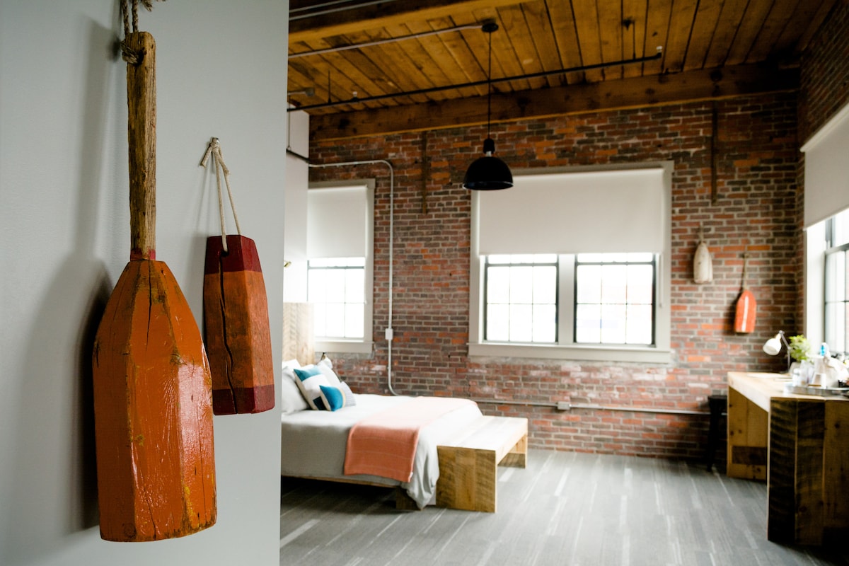 An interior view showcases warm wooden beams and exposed brick walls. A spacious bed is centered in the room, accompanied by colorful decorative pillows. Vintage-style wall decor, including red-painted paddle and hanging items, complements the light streaming through large windows.