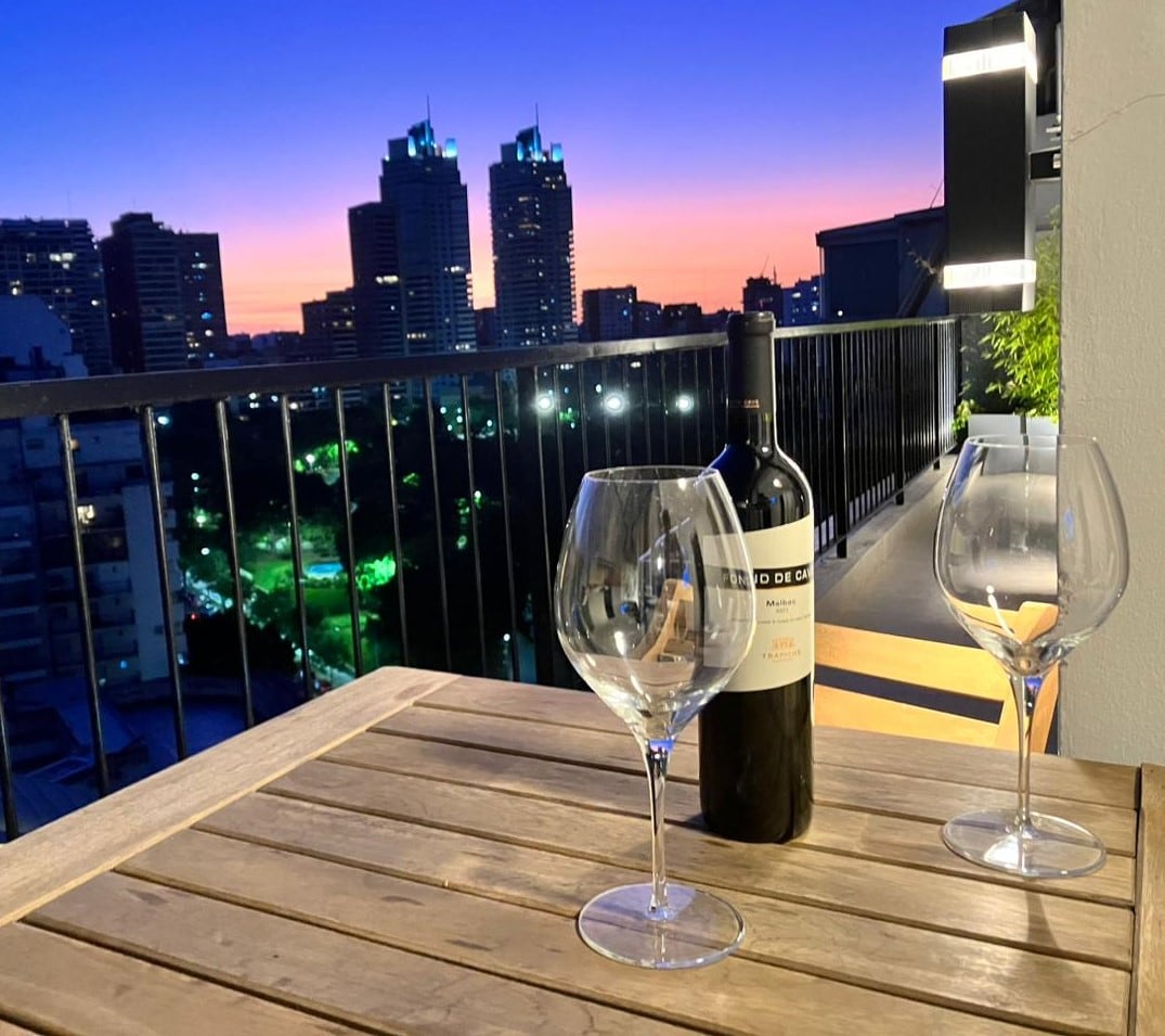A serene sunset view is framed from a balcony, showcasing a vibrant skyline with illuminated buildings. A wooden table is set with two large wine glasses and a bottle of red wine, inviting relaxation amidst the picturesque backdrop.