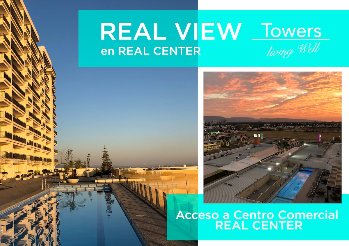 An inviting swimming pool stretches along the edge of a building, reflecting the warm evening light. In the background, a sunset creates a colorful sky over a shopping center, with various retail outlets visible below.