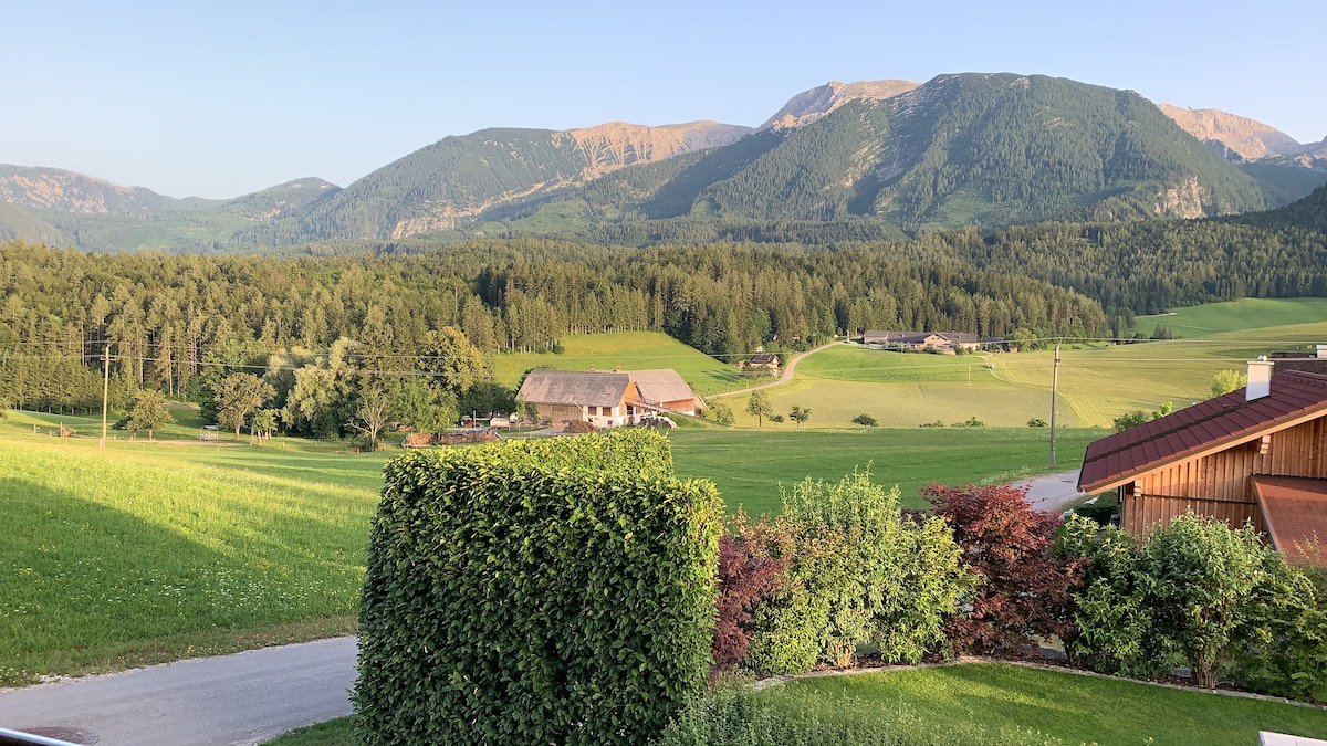 A panoramic view of lush green fields and gentle rolling hills is presented, with distant mountains in the background. The foreground features carefully trimmed hedges, while clusters of trees and farm buildings dot the landscape, emphasizing the serene natural setting.